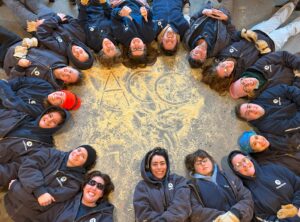 Members of AmeriCorps Cape Cod laying down in a circle, smiling, with "ACC" written in sawdust in the middle of the circle.