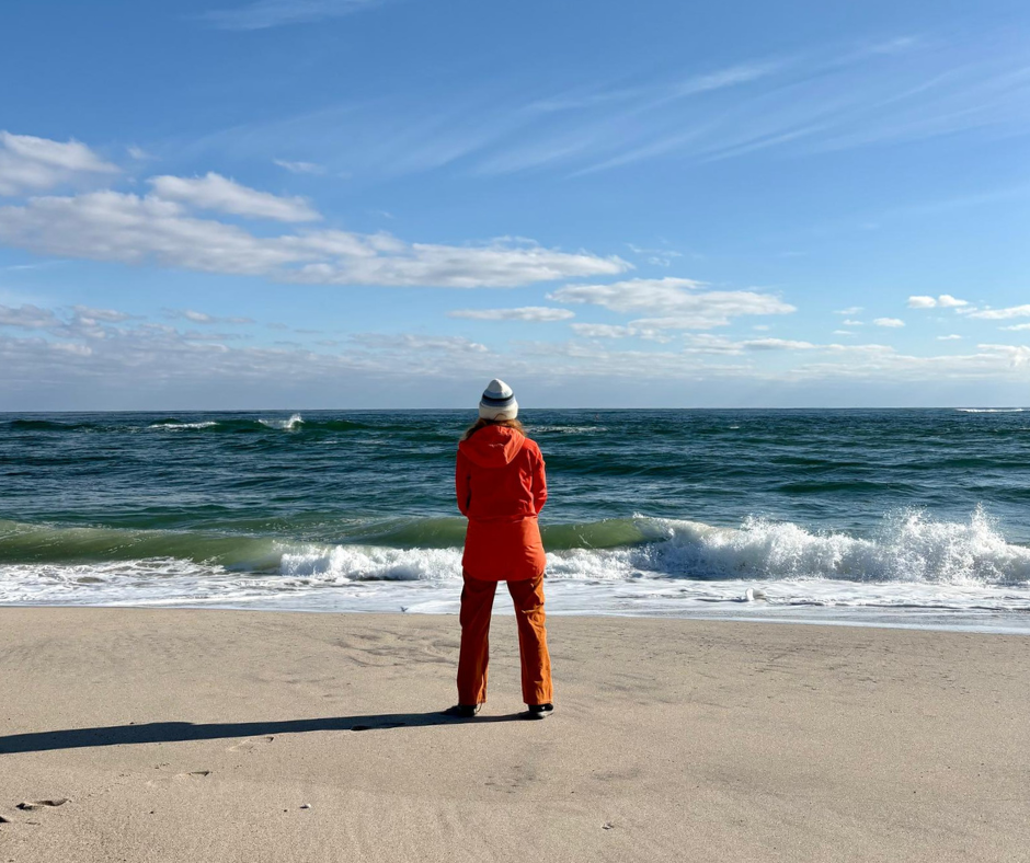 Person in red jacket and white hat standing alone on beach looking at ocean waves