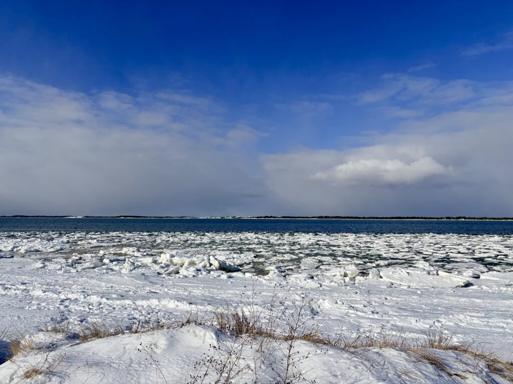 Beach shoreline partially covered with snow and ice, with frozen patches along the sand and water’s edge, under a blue sky with scattered clouds