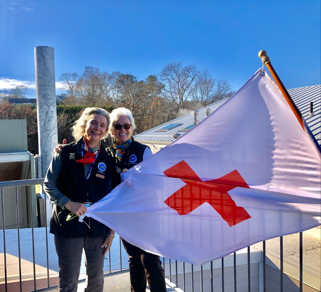 Two Barnstable County public health nurses smiling outdoors on a rooftop deck, holding a large white flag with a red cross, on a sunny winter day.