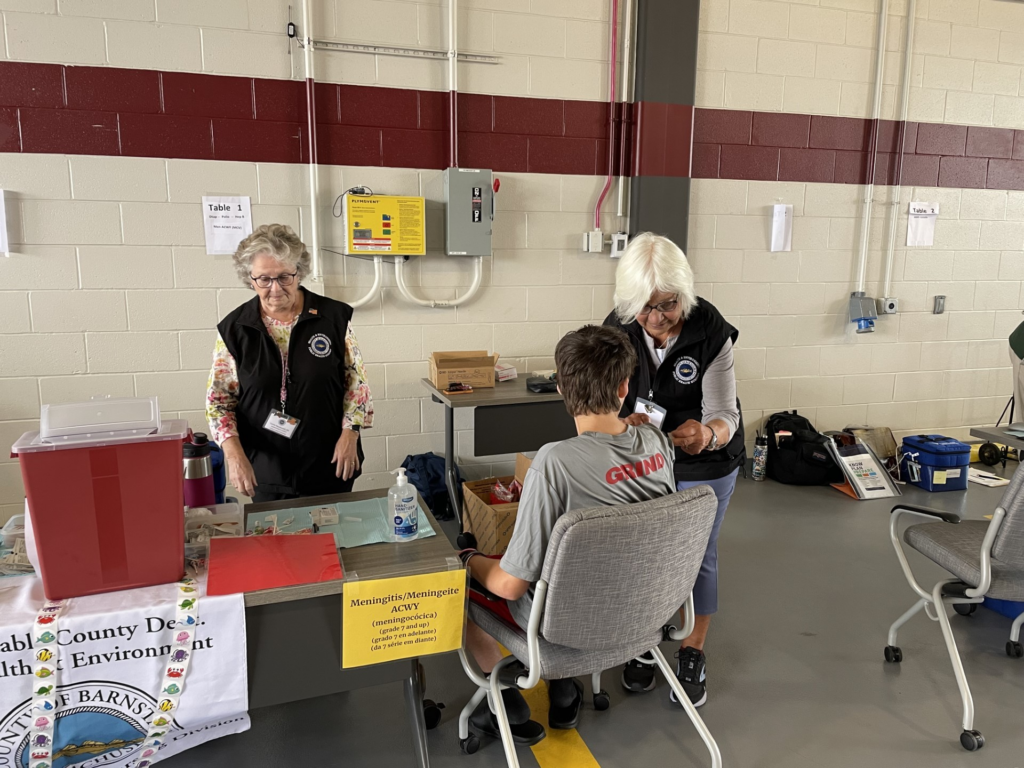 Two Barnstable County public health nurses administer a meningitis vaccine to a young person at a community immunization clinic, with a bilingual vaccine information sign visible on the table.