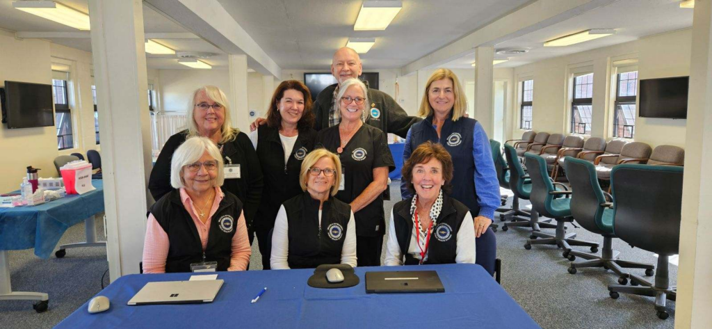 A group of eight Barnstable County Department of Health and Environment staff pose together at a community health event, wearing matching navy vests.