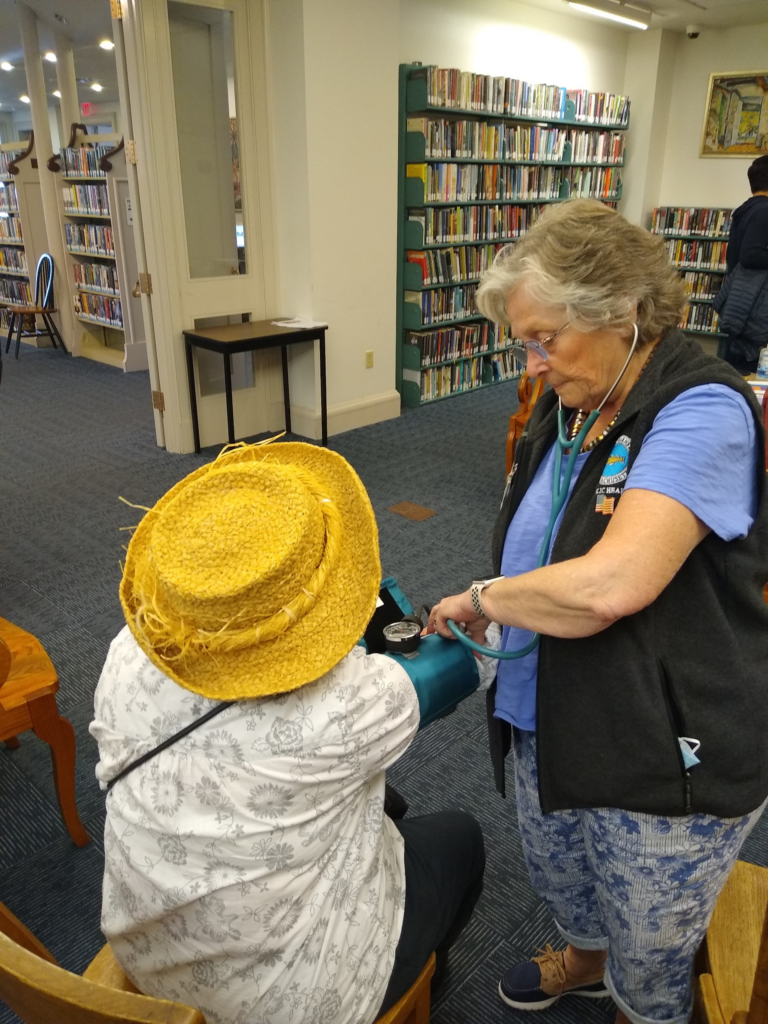 A Barnstable County public health nurse takes a community member's blood pressure at a public library health screening, with bookshelves visible in the background.