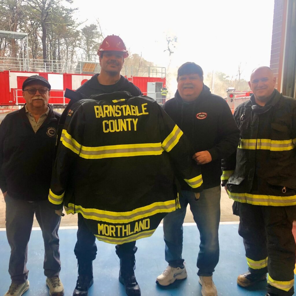 Group of four fire training professionals standing indoors holding a black firefighter jacket labeled “Barnstable County Morthland,” wearing a mix of protective gear and casual clothing, with fire training equipment visible in the background.
