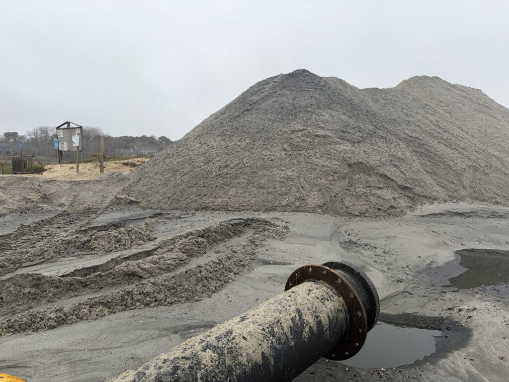 Dredge discharge pipe releasing sand onto the beach at Cockle Cove, forming large sand piles.