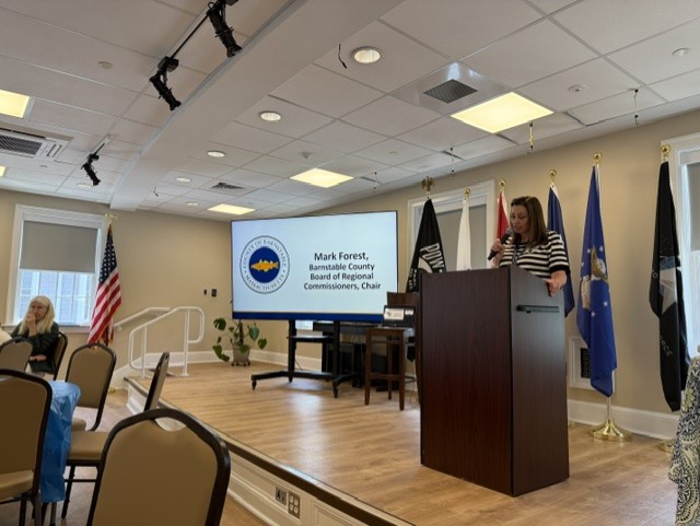 SHINE Program Manager Kristina Whiton-O'Brien speaks at the Cape Cod & Islands SHINE volunteer recognition event, with a presentation honoring volunteers displayed behind her.