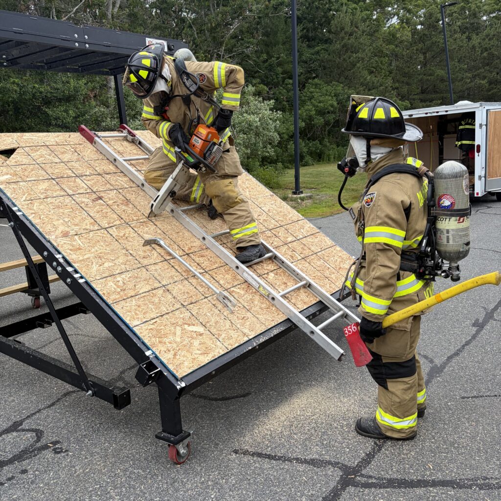 Two firefighters in full protective gear participate in a roof operations training exercise on a mobile roof prop. One firefighter kneels on a ladder positioned on a simulated pitched roof while using a chainsaw to cut through the roof surface, as sawdust sprays from the cut. A second firefighter stands below with a fire axe, observing and assisting. The training setup is located outdoors, with fire department equipment and trees visible in the background.