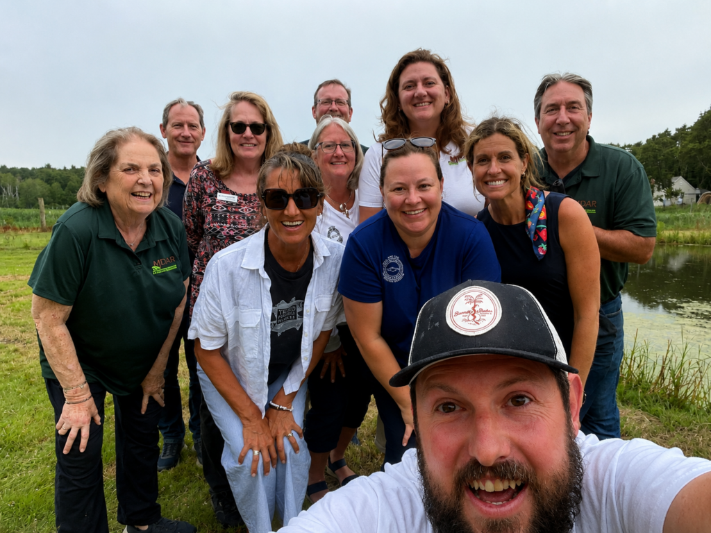 Smiling group selfie of Massachusetts Department of Agricultural Resources (MDAR) staff and Cape Cod Cooperative Extension staff during a farm tour at Hillside Farm in Truro, with fields and a pond visible behind the group.