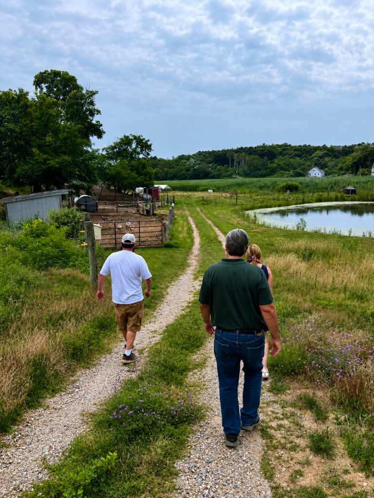 Participants walk down a gravel farm road at Hillside Farm in Truro toward fields, livestock pens, and a pond during a guided tour of the farm. The surrounding landscape includes pasture, crops, and wooded hills beneath a cloudy sky.