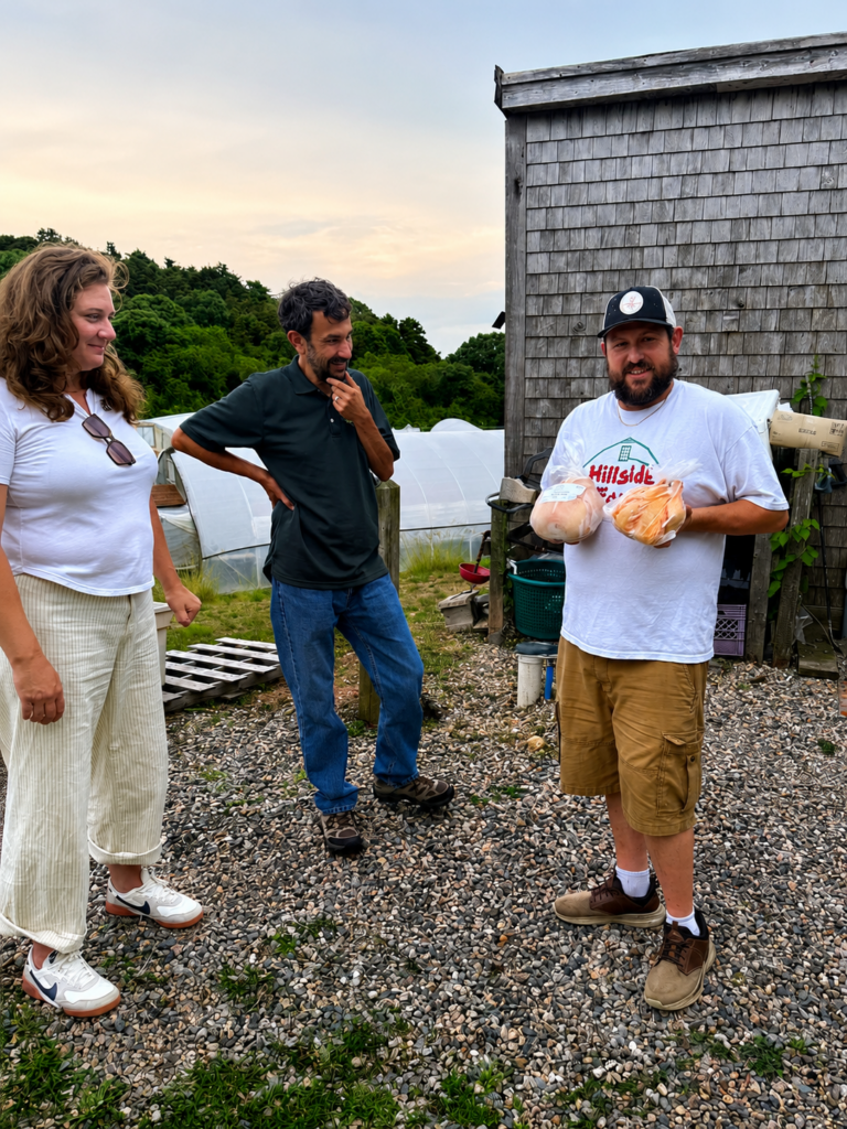 At Hillside Farm in Truro, Drew Locke holds two locally raised chickens while speaking with visiting Massachusetts Department of Agricultural Resources (MDAR) staff outside the farm buildings. Two attendees listen nearby, with greenhouses visible in the background.