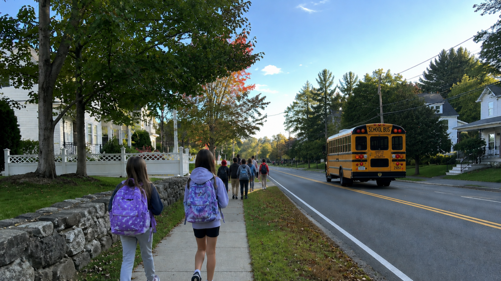 Children with backpacks walk along a tree-lined sidewalk on a sunny early-fall morning as a yellow school bus travels down the road beside them, in a New England neighborhood.