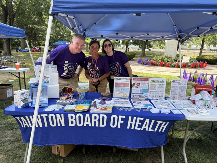 Three people in matching purple Taunton Board of Health T-shirts stand smiling behind an outreach table at an outdoor event. The blue tablecloth reads 'TAUNTON BOARD OF HEALTH' in large white letters, and the table is covered with health education flyers, brochures, giveaways, and an emergency kit. A blue pop-up tent shades the booth, with a grassy park and rows of small flags visible in the background. Cassidy O'Hara is on the right.