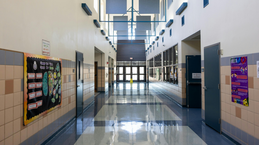 "An empty, sunlit school hallway with polished tile floors, rows of classroom doors, and bulletin boards on the walls, leading toward glass entrance doors at the far end."