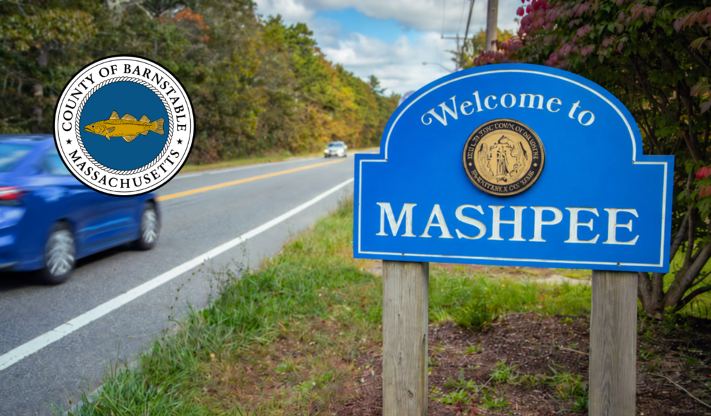 A blue “Welcome to Mashpee” roadside sign stands beside a tree-lined road, with a blue car passing in the foreground. The Barnstable County, Massachusetts seal is overlaid in the upper left corner.