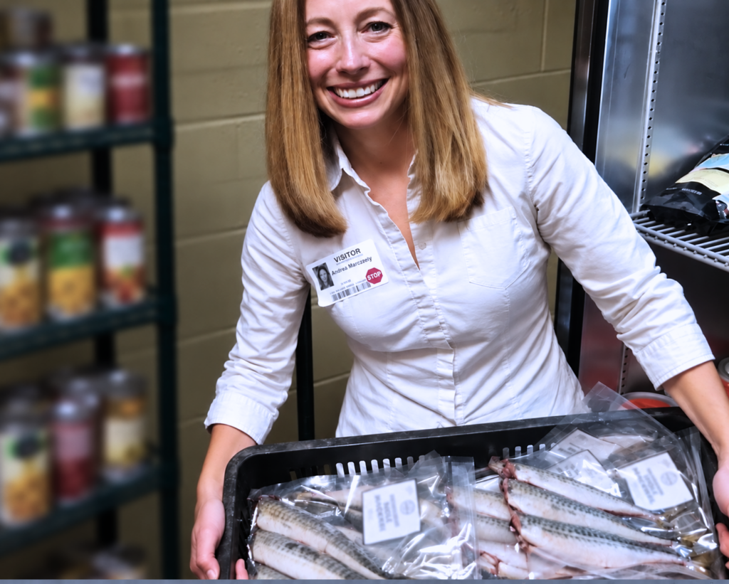 Andrea Scarpellini smiles while holding a crate of packaged frozen fish destined for Cape Cod food pantries, with shelves of canned food beside her.
