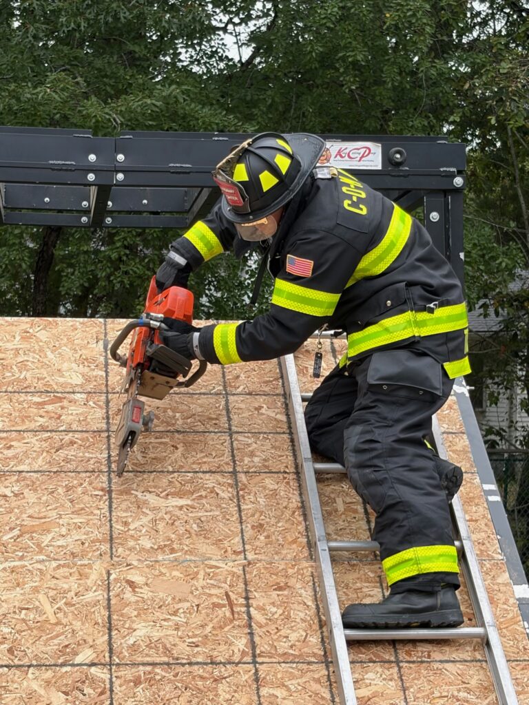 Firefighter in protective gear and a black helmet uses a chainsaw to cut a wooden roof training panel while positioned on a ladder.